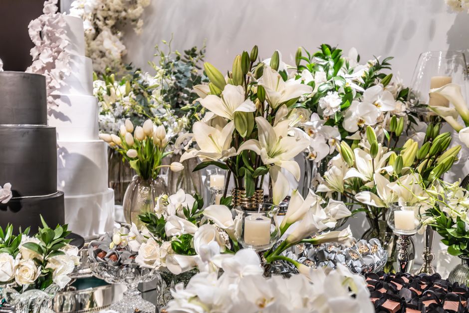 A luxurious floral arrangement featuring white lilies and white orchids, complemented by small white roses and greenery, is displayed on a reflective surface. The bouquet is surrounded by glass candle
