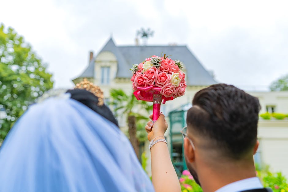 A person holding a round bouquet of fresh pink and white roses, with small white filler flowers, wrapped with pink ribbon, positioned outdoors in front of a historic house with a slate roof and dormer