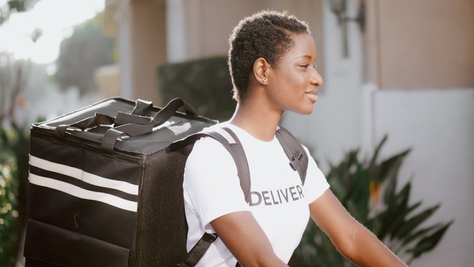 A young woman with short, curly hair and medium skin tone, wearing a white T-shirt with 'DELIVER' printed on it, is standing outdoors in natural light. She has a black backpack with white stripes on h