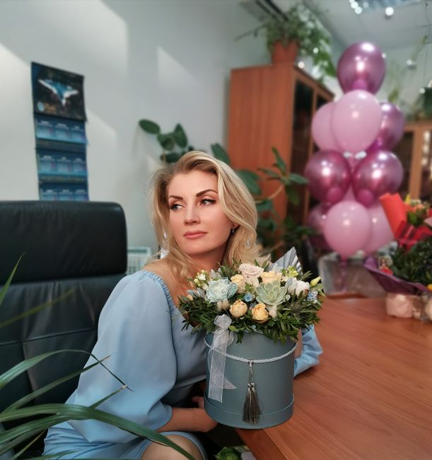 A woman with blonde hair and a light blue blouse is seated at a wooden desk in a floral shop, holding a round, elegant floral arrangement in a light blue hat box decorated with a gray ribbon and tasse