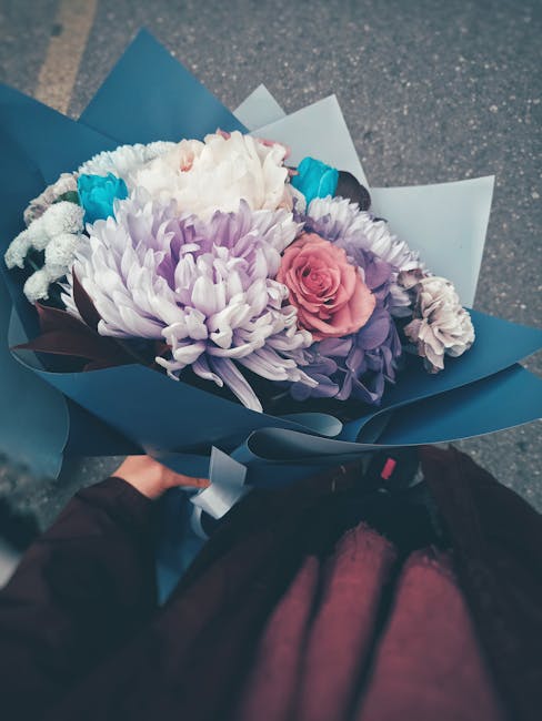 A person holding a floral bouquet wrapped in dark blue and light blue paper, featuring a mix of fresh flowers including a large white chrysanthemum, a soft pink rose, and smaller purple and blue bloom