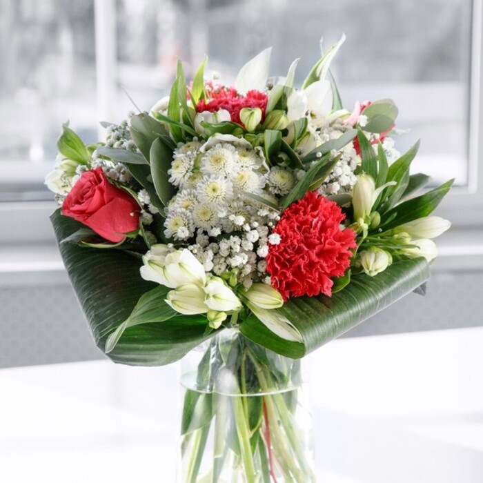 In this image, a meticulously crafted red and white bouquet rises with quiet confidence from a clear cylindrical glass vase, the water inside magnifying the tidy network of green stems like a window into the arrangement's foundation. At the heart of the design, deep red roses stand slightly taller, their velvety petals gently unfolding, as if caught at the moment when a romantic gesture is first revealed. Nestled close to them is a large crimson carnation, its intricate, frilled petals packed tightly together, offering a tactile counterpoint to the smooth curves of the roses. Around these rich reds, a constellation of white blooms creates balance and serenity: white alstroemeria with slender petals and fine inner speckles, petite chrysanthemum-like blossoms, and the soft hint of creamy tones that prevent the whites from feeling stark. Several elegant white lily buds are still closed, their elongated forms suggesting grace and the promise of continued beauty in the days ahead. Wisps of gypsophila-baby's breath-drift between the larger flowers, their tiny, cloudlike clusters giving the whole arrangement a softly romantic, almost weightless finish. Encircling the bouquet, lush greenery in multiple shades builds structure and depth. Broad, dark green foliage is artfully curled and wrapped around the base, forming a smooth, sculpted collar, while more slender, eucalyptus-style leaves and pointed stems weave upward, adding movement and a natural garden feel reminiscent of hedgerows and green corners around Beddington. The bouquet rests on a clean white surface under gentle, even daylight, with the background blurred into a soft haze that suggests a bright room or a window facing a quiet residential lane. The overall impression is of refined, modern romance-an ideal choice for a dining table, a special anniversary surprise, or a warm thank-you delivered to a friend or colleague living or working in Beddington.