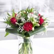 A luxurious red and white bouquet stands proudly in a clear cylindrical glass vase, its waterline revealing neatly trimmed green stems that speak of expert care and same-day freshness in Beddington. Deep, velvety red roses rise at the heart of the arrangement, each petal just beginning to unfurl, giving the sense of romance caught mid-breath. At the front, a large crimson carnation, ruffled and richly layered, draws the eye with its intricate texture, like fabric gathered and pleated by hand. Around these passionate reds, clusters of pure white and creamy blooms glow softly: white alstroemeria with slender petals and speckled throats, tight white lily buds poised to open over the coming days, and airy clouds of delicate baby's breath that seem to float between the larger flowers. Lush greenery frames the bouquet, with broad, glossy dark green leaves elegantly curled and wrapped around the base like a collar, while slimmer eucalyptus-like foliage and pointed leaves weave upward, adding depth, shape, and several tones of green. The arrangement rests on a pristine white surface, possibly a tabletop in a bright Beddington home, with a softly blurred, light-filled background that suggests a window looking out toward a quiet residential street or a leafy corner near Beddington Park. Gentle, even daylight touches every petal, making the reds appear rich and saturated and the whites clean and luminous. The overall impression is one of polished celebration and heartfelt warmth, ideal as a centrepiece for an anniversary dinner, a spring birthday, or a thoughtful delivery to brighten a loved one's office near the centre of Beddington, carrying a message of admiration, joy, and enduring affection.