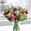 A luxurious red and white bouquet sits proudly in a clear glass vase on a bright white tabletop, the kind of elegant arrangement you might find gracing a dining room near Beddington Park on a crisp morning. Deep crimson roses, some fully unfurled with velvety petals and others still in tight, promising buds, cluster at the heart of the design, giving the whole display a romantic, almost timeless feel. Nestled among them are rich red carnations, their ruffled, full blooms adding volume and a sense of classic charm, like something chosen for an anniversary dinner or a heartfelt thank-you. Threaded between the red flowers, white alstroemeria lilies offer a gentle contrast; many of their buds are still closed or just beginning to open, showing off slender, trumpet-shaped petals tipped delicately with green. Tiny sprays of white gypsophila-baby's breath-are scattered generously throughout, creating a soft, airy halo that lightens the bold reds and ties the colour palette together. Around the base and weaving through the centre, lush green foliage forms a structured frame: broad, glossy leaves create a collar at the bottom, while narrower, more flexible greenery snakes up between the blooms, giving movement and depth. The stems are clearly visible through the vase, standing in clean, fresh water that subtly suggests same-day preparation and delivery from a local Beddington florist. Behind the arrangement, a large window lets in diffused natural light, and the lightly blurred, almost monochrome outdoor view ensures that the reds, whites, and greens are what truly capture the eye, making this bouquet ideal for celebrating milestones, romantic gestures, or simply brightening a home along Beddington Lane or in the quiet streets nearby.